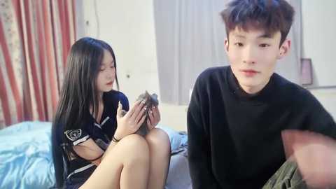 Media: Video of an Asian woman with long black hair, wearing a navy uniform, holding a small animal, sitting on a bed; an Asian man with short dark hair, wearing a black sweater, looking at her.