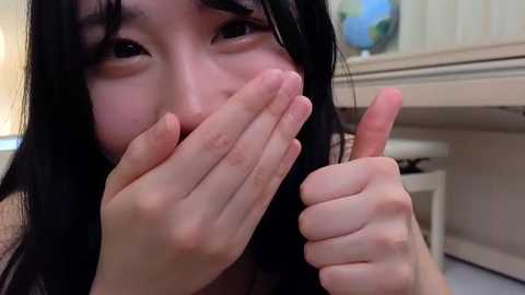 Media: A close-up video of an Asian woman with long black hair, giving a thumbs-up while playfully hiding her nose with her fingers. She has a light skin tone and is indoors, with a beige kitchen background visible.