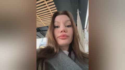 Media: A video of a young woman with fair skin, long brown hair, and light makeup, wearing a grey sweater, indoors with a wooden ceiling and blurred background.