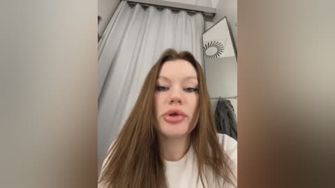 Media: Video of a fair-skinned, young Caucasian woman with long brown hair, wearing a white t-shirt, sticking out her tongue. Background includes a white shower curtain, towel rack, and ceiling fan.