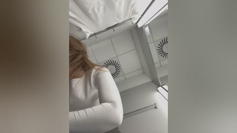 Media: Video of a woman in a white long-sleeve shirt, viewed from behind, standing in a white-tiled shower with a ceiling fan and a towel rack.