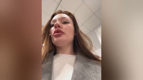 Media: Video of a young woman with fair skin, long brown hair, and a slight smile, wearing a grey cardigan over a white shirt, standing in a tiled office with a white ceiling.