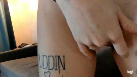 Media: Video of a fair-skinned person's leg with a tattoo of \"N\" and a heart, holding a phone, indoors, blurry background.