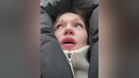 Media: Video of a young girl with fair skin, wearing a gray puffy jacket and a white ribbed sweater, lying on her back, looking up with wide eyes and slightly open mouth, surrounded by blurred, gray, textured fabric.