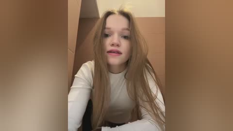 Media: A video of a young Caucasian woman with long, straight brown hair, wearing a white long-sleeved top, peering out from a cardboard box. The background is a beige wall.