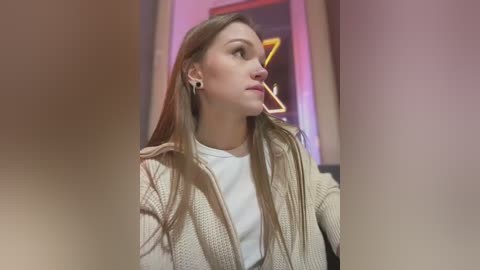 Media: A video of a young Caucasian woman with light brown hair and fair skin, wearing a beige knit cardigan over a white shirt. She has black ear gauges and a cigarette in her mouth, looking thoughtful.