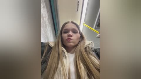 Media: A video of a young woman with long, straight blonde hair, wearing a white sweater and black jacket, sitting on a bus, looking straight at the camera.