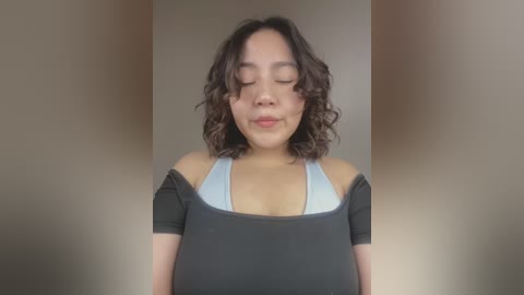 Media: Video of a young, light-skinned Asian woman with wavy brown hair, wearing a black off-shoulder top revealing a light blue bra, smiling softly with closed eyes.