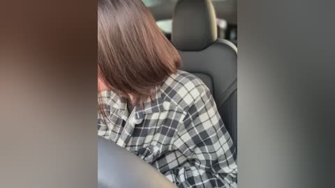 Media: Video of a person with shoulder-length brown hair, wearing a black and white plaid shirt, sitting in the back seat of a car with black leather upholstery.