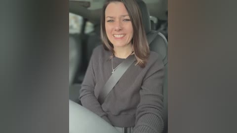 Media: Video of a smiling Caucasian woman with shoulder-length brown hair, wearing a gray sweater, seated in a car with a gray seatbelt.