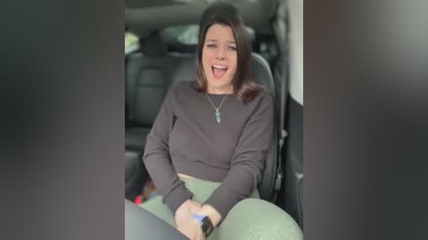 Media: Video of a woman with shoulder-length brown hair, wearing a dark sweater, sitting in a car, laughing, holding a blue device, surrounded by blurred car interior.