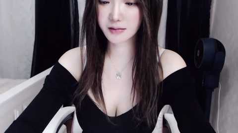 Media: Video of a young Asian woman with long brown hair and pale skin, wearing a black off-shoulder top, seated in a dimly lit room with a white bathtub and a black speaker.