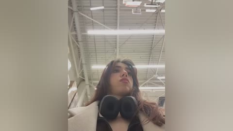 Media: A video of a woman with long brown hair and fair skin, wearing black headphones and a beige coat, taken from a low angle inside a warehouse with a metal ceiling and fluorescent lighting.