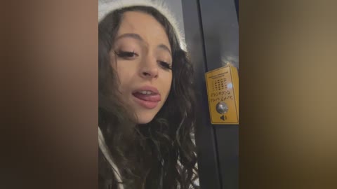 Video of a young woman with long, curly dark hair, wearing a white hoodie, leaning against a yellow metal door with a \"Push to Open\" sign, indoors, dim lighting.