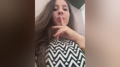 Media: Video of a young woman with long, wavy brown hair, wearing a black-and-white zigzag patterned top, holding a finger to her lips in a playful, secretive gesture.