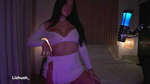 Media: Video of a woman with long, straight black hair wearing a white, off-shoulder bralette and high-waisted panties, standing in a dimly lit bedroom with a bed and a glowing blue lamp in the background.