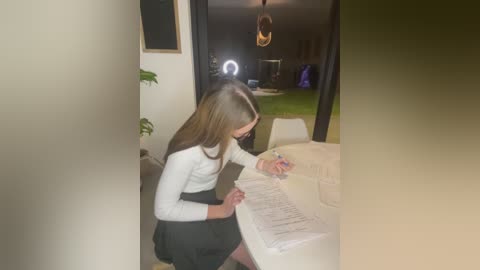 Media: Video of a young woman with long brown hair, wearing a white top and black skirt, seated at a white table, reading a magazine, in a dimly lit room with a green plant and a window in the background.