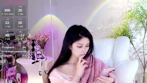 Media: Video of an Asian woman with long black hair, wearing a pink off-shoulder top, texting on her phone, in a bright, minimalist room with a white couch and greenery, with a rainbow effect in the background.