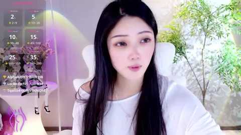 Media: A video of an East Asian woman with long black hair, fair skin, and light makeup, sitting in a white chair, wearing a white top. The background features a green plant and a calendar display.