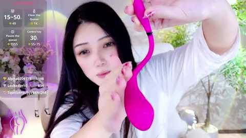 Media: Video of an Asian woman with long black hair, wearing a white top, holding a bright pink bra, against a green background.