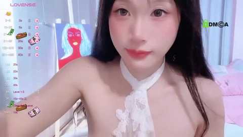 Media: A video of an East Asian woman with long black hair and fair skin, wearing a white lace halter top, in a bedroom with pink bedding and a colorful wall painting.