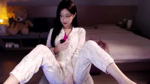 Media: Video of an East Asian woman with long black hair, fair skin, and a slender build, wearing white pajamas, sitting on a man's lap, in a dimly-lit bedroom with a teddy bear, candles, and a bed in the background.