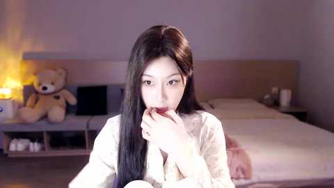 Media: Video of an Asian woman with long black hair, wearing a white robe, eating a donut, in a dimly lit bedroom with a teddy bear, a bed, and a lamp.