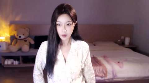 Media: Video of an East Asian woman with long black hair in a white floral-patterned robe, standing in a softly lit bedroom with a teddy bear, a warm lamp, and a neatly made bed.