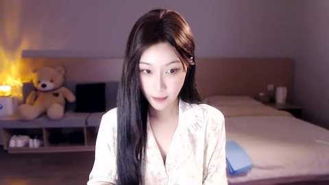 Media: Video of a young Asian woman with long black hair, fair skin, and delicate features, wearing a white floral-patterned nightgown, sitting on a bed in a dimly lit bedroom with a teddy bear and a lamp.