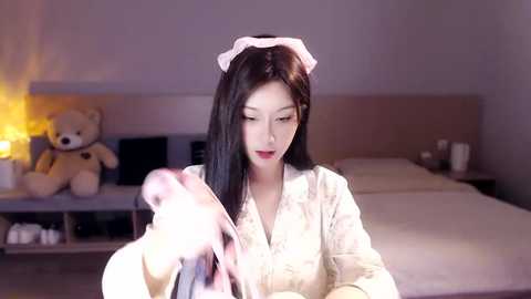 Media: Video of an Asian woman with long black hair, wearing a maid outfit, holding a pink feather duster in a modern bedroom with a beige bed, teddy bear, and a lamp.