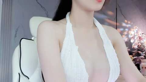 Media: A video of a fair-skinned woman with long black hair, wearing a white halterneck swimsuit, sitting on a white gaming chair in a dimly lit room with a marble wall and Christmas decorations in the background.