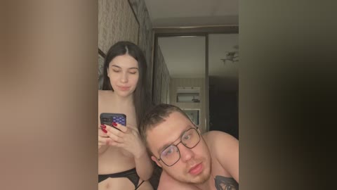 Media: A video of a young couple taking a mirror selfie in a dimly lit bathroom. The woman, with long dark hair, is topless, while the man, with glasses and tattoos, is shirtless.