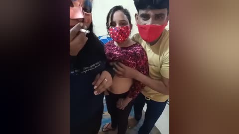 Media: Video of a young woman wearing a pink patterned crop top and black pants, with a red mask, surrounded by three men in casual clothing, indoors.