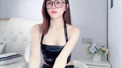 Media: Video of a slender, fair-skinned woman with long, straight red hair, wearing black-rimmed glasses and a black latex halter top, seated on a white, tufted chair in a minimalist, brightly lit bedroom.