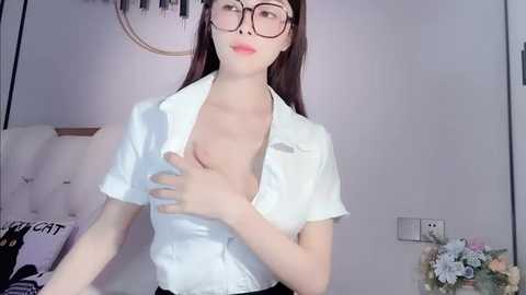 Media: A video of an East Asian woman with fair skin and long, straight brown hair, wearing a white blouse unbuttoned to reveal her medium-sized breasts, black-framed glasses, and a hint of makeup. She stands in a minimalist bedroom with a bed, floral arrangement, and wall art.