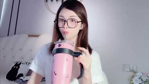 Media: Video of a young, fair-skinned woman with glasses, drinking from a large, pink insulated tumbler with a straw, wearing a white t-shirt, in a white room with a bed and a bouquet of flowers.