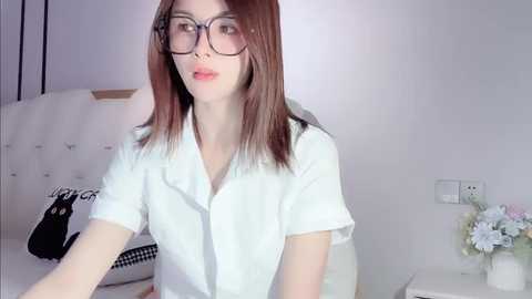 Media: Video of a slender, fair-skinned woman with straight, shoulder-length brown hair, wearing glasses and a white blouse, sitting in a minimalist, modern bedroom with a white bed, floral arrangement, and electrical outlets visible.