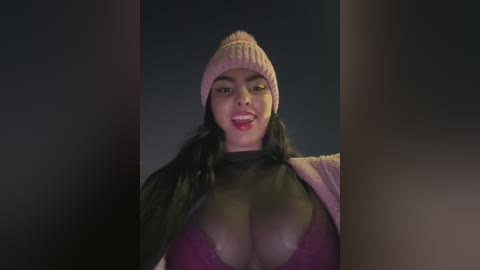 Media: Video of a smiling, curvaceous Black woman with long black hair, wearing a pink beanie, black turtleneck, and a sheer pink top revealing her large breasts. She stands in front of a dark, blurry background.