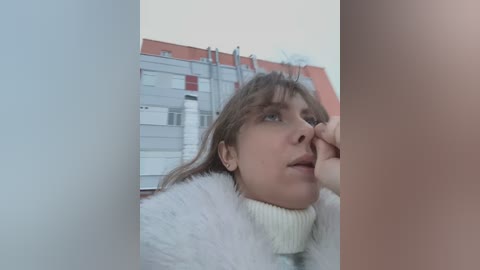 Media: Video of a young woman with light skin, light brown hair, and a white fur coat, standing thoughtfully by a building with red-tiled roofs and metal railings.