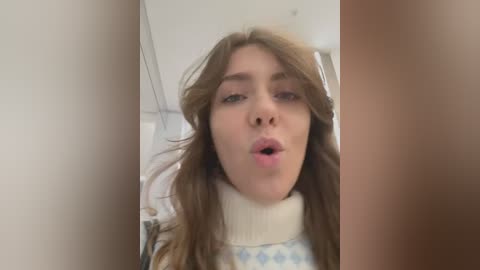 Media: Video of a young Caucasian woman with long, wavy brown hair, wearing a white turtleneck sweater, pouting with lips slightly parted. Background is blurry, featuring a light-colored wall.