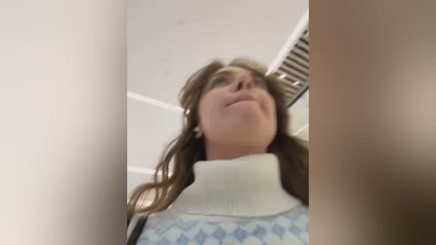 Media: Video of a young woman with long brown hair, wearing a white turtleneck and blue diamond-patterned sweater, looking up at a ceiling fan in a dimly lit room.