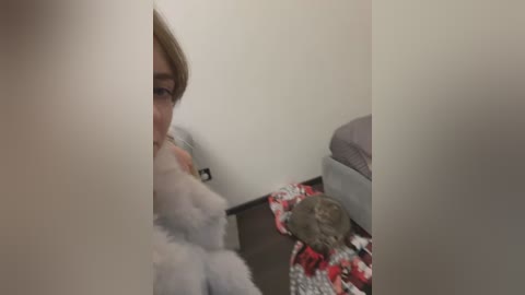 Media: Video of a young woman with light skin and straight, light brown hair, wearing a white dress, partially obscured by a fluffy white blanket. She sits on a dark wooden floor next to a gray bed with red and white patterned blankets. A brown cat lies beside her.