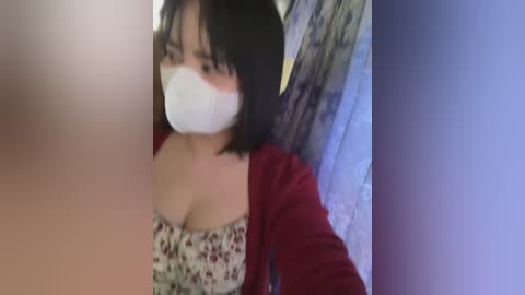 Media: Video of an Asian woman with short black hair, wearing a floral top, red cardigan, and white face mask, indoors, blurred background.