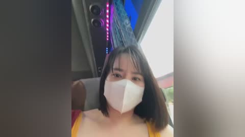 Media: Video of an Asian woman with shoulder-length black hair, wearing a yellow tank top and a white face mask, sitting in a dimly lit vehicle with blue and pink neon lights in the background.