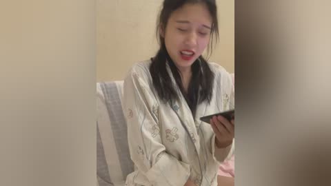 Media: Video of an Asian woman with long black hair in pajamas, looking down at a smartphone, sitting on a striped bed in a dimly lit room.