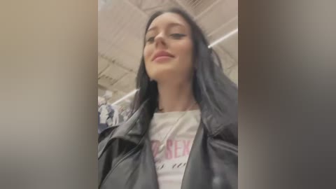 Media: Video of a young woman with long black hair, fair skin, and a confident smile, wearing a black leather jacket over a white graphic tee, taken in a dimly lit, industrial-looking room with exposed ceiling panels.