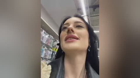 Media: A video of a smiling woman with fair skin, black hair, and wearing a black leather jacket, taken indoors near a brightly lit store shelf.