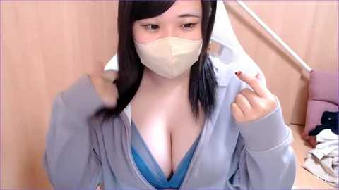 Media: Video of an East Asian woman with light skin, wearing a gray hoodie, exposing a blue bra revealing ample cleavage, a beige face mask, and straight black hair, against a wooden backdrop with a pink chair.