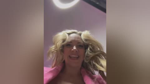 Media: Video of a smiling woman with platinum blonde hair styled into voluminous curls, wearing a pink robe. The background is a blurred, pink-toned indoor setting with a ceiling light.