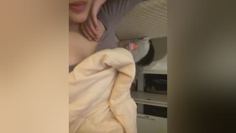 Media: Video of a woman lying in bed, partially covered by a beige blanket, with a grey wall and a white dresser in the background.